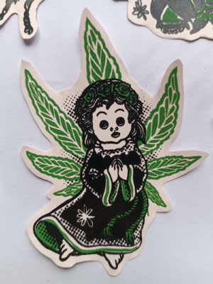 Adesivo com desenho estilizado de anjo criança europeu, com uma folha de cannabis aberta atraz de suas costas, parecendo asas. Arte em duas cores variáveis (preto +cor)