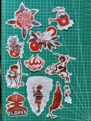 11 adesivos variados sobre fundo verde quadriculado. Curupiras, Saci-Pererê, explosão em floreira, regador com planta crescendo dentro, canário em galho de laranjeira, flor de cannabis, pandeiro em chamas, artesanato de palha trançada parecendo uma espiga de milho, e uma bergamota semi-aberta em formato de flor ou estrela. Desenhos em marrom escuro e vermelho, fundo dos adesivos offwhite.